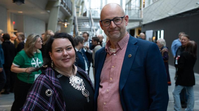 dame med kofte og mann med dress i en hall med mange mennesker bak