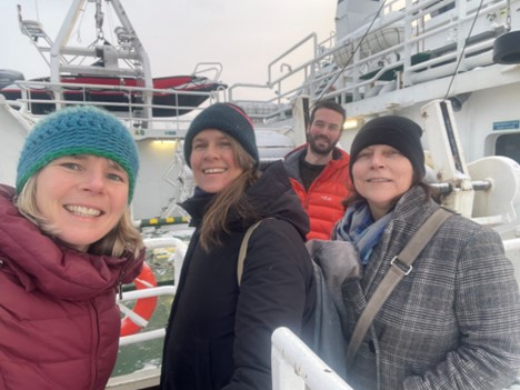 Picture1.jpg Fire personer ombord på et stort skip. I forgrunnen fra venstre står en blond kvinne i rød boblejakke og blå strikket lue, en kvinne med brunt hårr og svart jakke, samt en kvinne i rutete jakke og svart lue. Bak dem står en mann med mørkt hår og skjegg i orange boblejakke. I bakgrunnen ser vi at de er på et skip, med hvite rekkverk og vegger, livbåt, lettbåt og deler av maskineri.