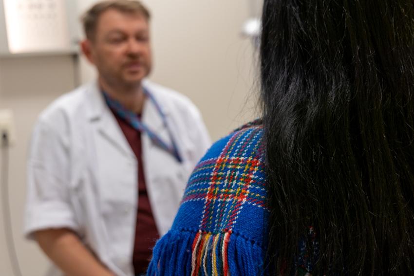 Ein mann i legefrakk ser på ei kvinne på kontoret. Kvinna har ryggen til og har på samisk sjal.