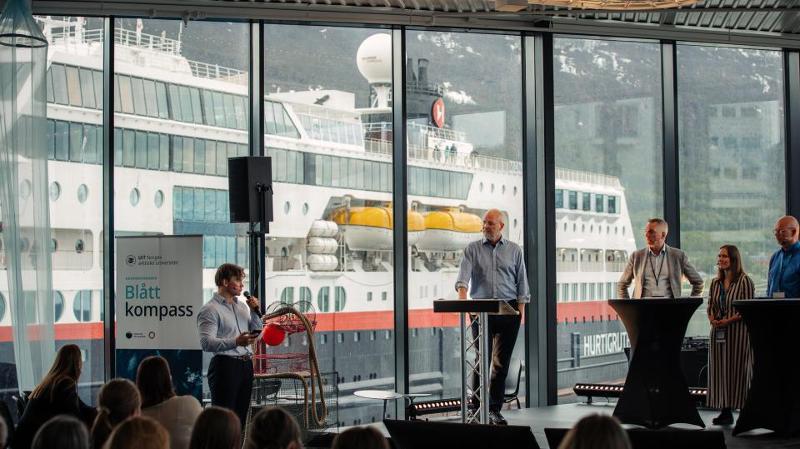 mennesker som st&aring;r p&aring; en scene og har debatt, stort vindu i bakgrunnen der man kan se en stor b&aring;t komme til kai