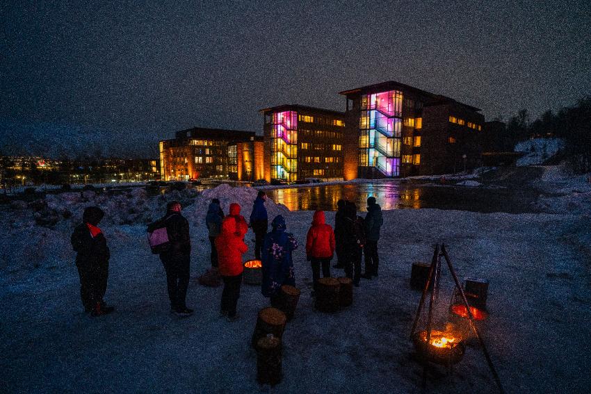 Bildet er tatt i Midtparsellen på campus i Breivika- I forgrunnen er en bålpanne med flammer og mennesker som ser på den. I bakgrunnen sees Teorifagsbyggene som er lyssatt. Det er mørkt ute.