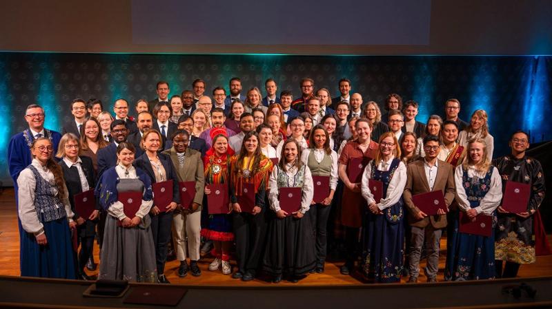 mange festkledde mennesker som st&aring;r sammen p&aring; en scene med diplom