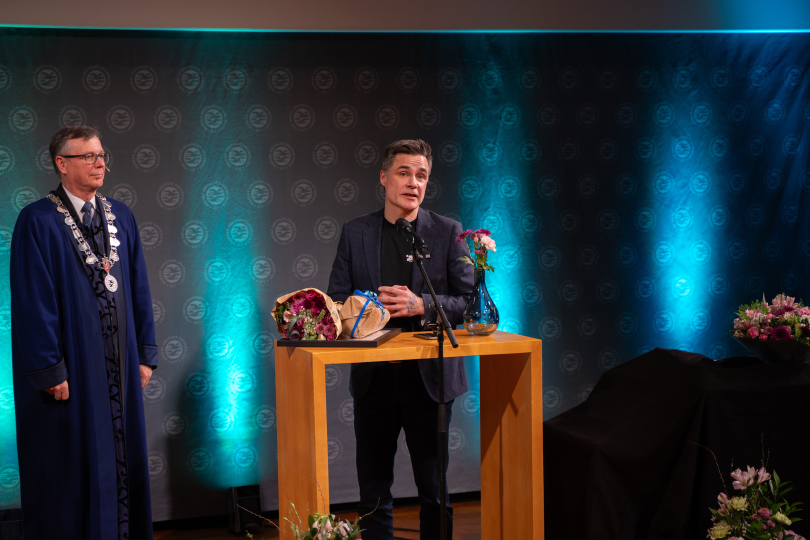 En mann mottar diplom, blomster og gave i staselig og feststemt universitetsauditorium. 