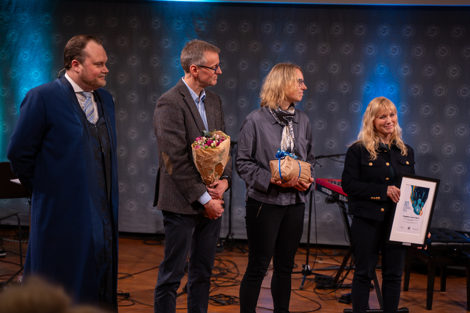 To damer og en mann mottar diplom, blomster og gave i staselig og feststemt universitetsauditorium. En rektor til venstre for dem. 