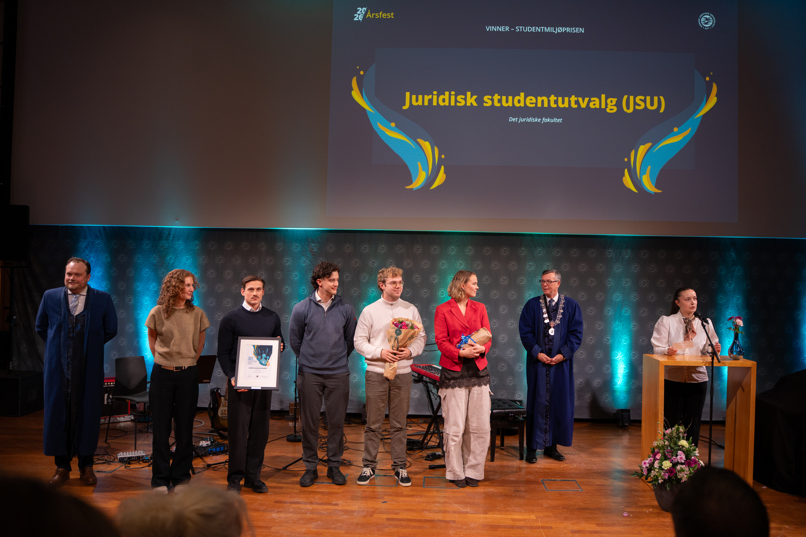 En gruppe entusiastiske unge studenter og ansatte mottar diplom, blomster og gave i staselig og feststemt universitetsauditorium. 