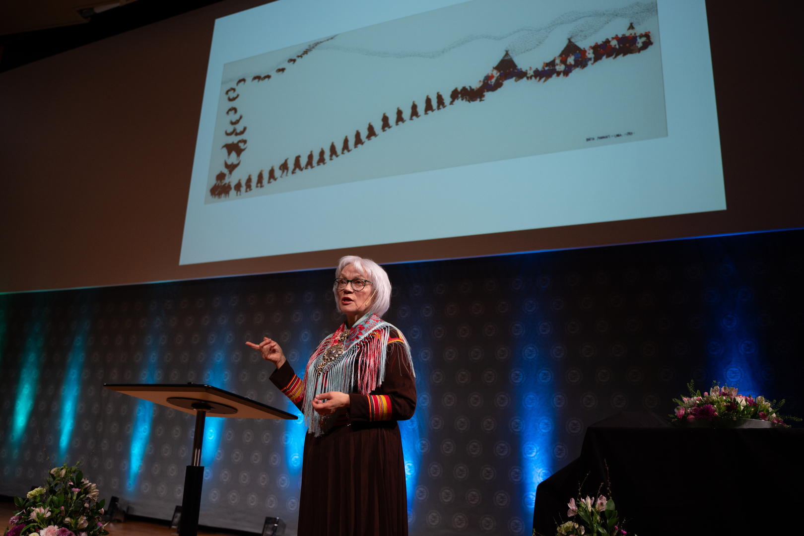dame med kofte som st&aring;r p&aring; en scene med kunstverk i bakgrunnen