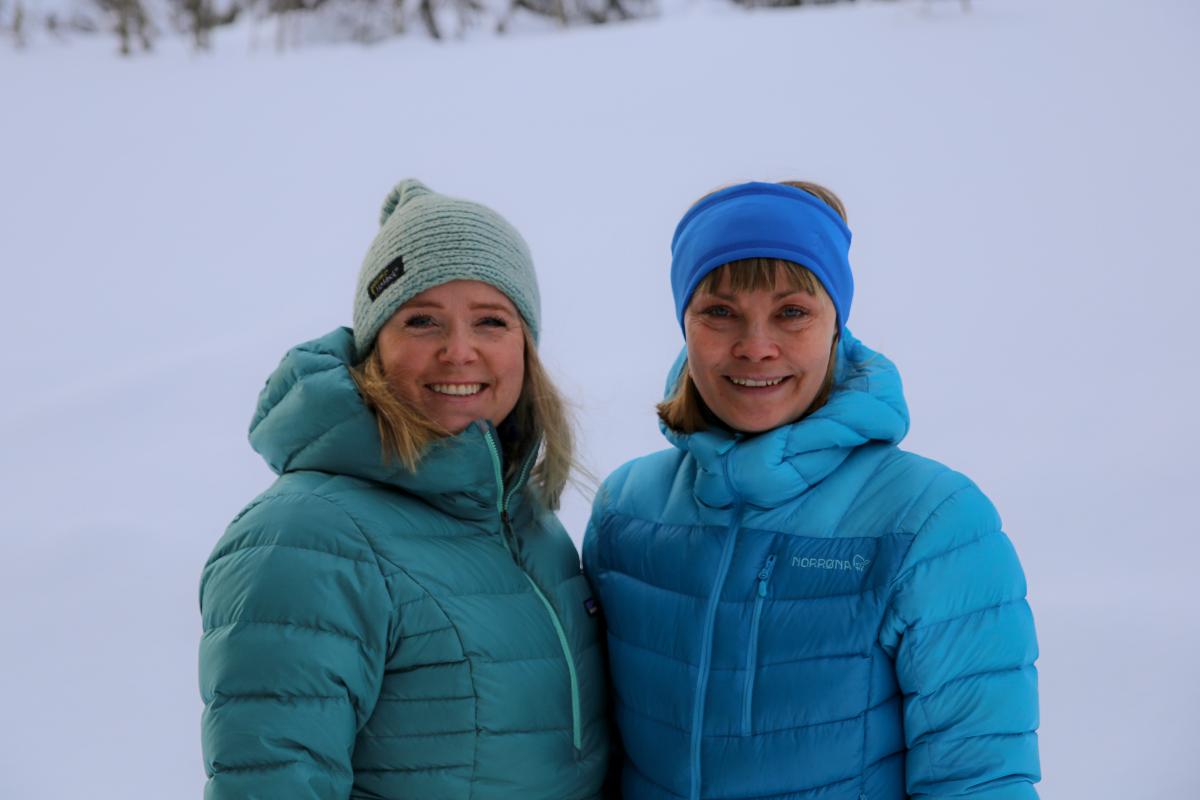 to kvinner, side om side, smiler, sn&oslash; i bakgrunnen, kvinne til venstre har blondt, skulderlangt h&aring;r, bl&aring;gr&oslash;nn jakke, sj&oslash;gr&oslash;nn lue, kvinne til h&oslash;yre har kort blondt h&aring;r, bl&aring; jakke og panneb&aring;nd. 