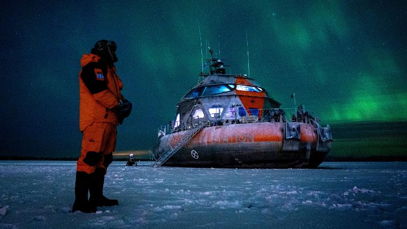 en person i overlevelsesdrakt st&aring;r p&aring; isen ved et rundt og oransje fart&oslash;y. nordlys i polarnatten over dem.