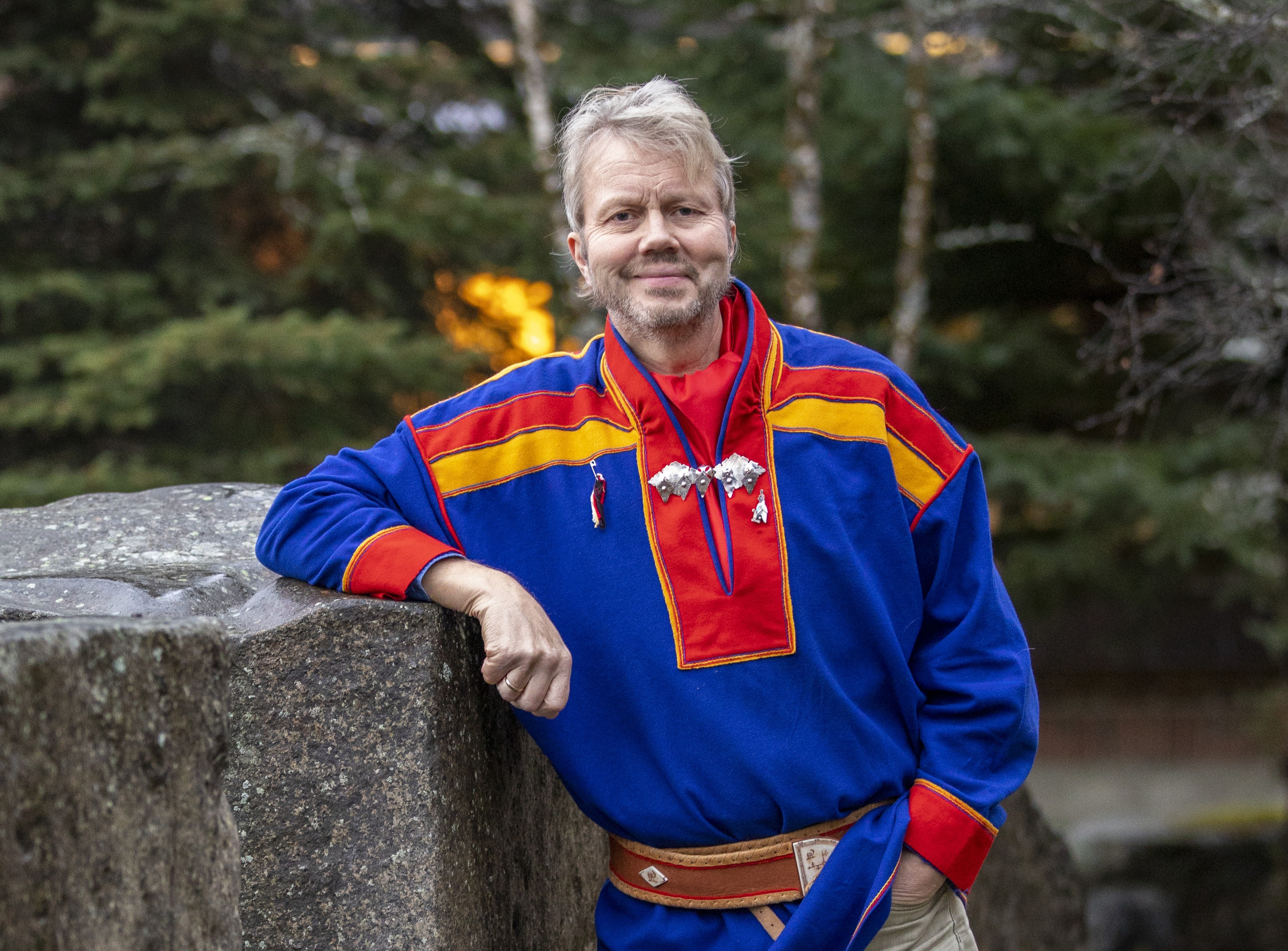 mann, i f&oslash;rt samisk drakt, venstre h&aring;nd i bukselomme, h&oslash;yre albue hviler p&aring; en h&oslash;yde, natur i bakgrunn, er et portrettbilde, mann ser inn i kamera