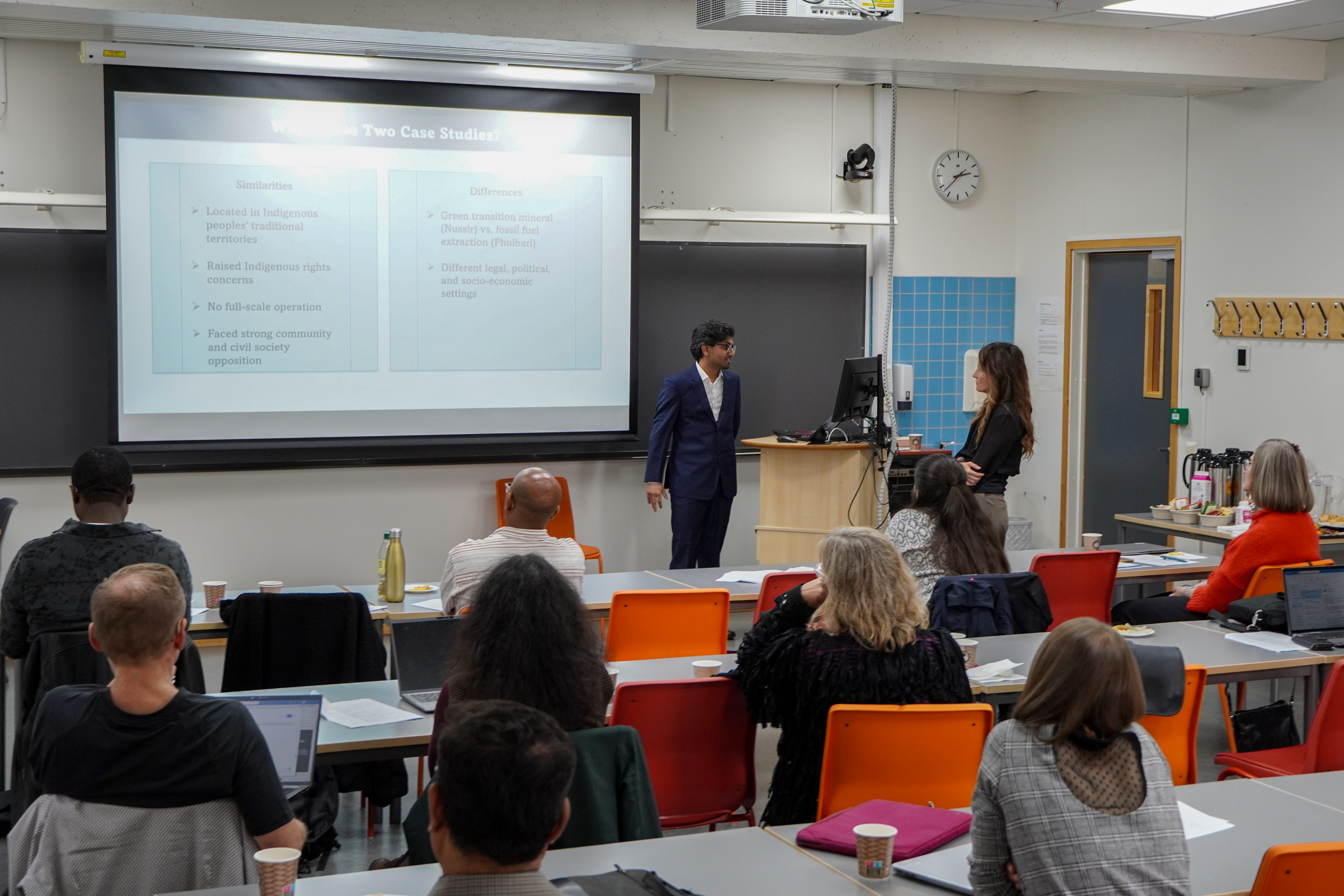 DSC02281.jpg Man, in suit, short dark hair, glasses, presenting in a classroom, screen in front of him, presentation in the back.
