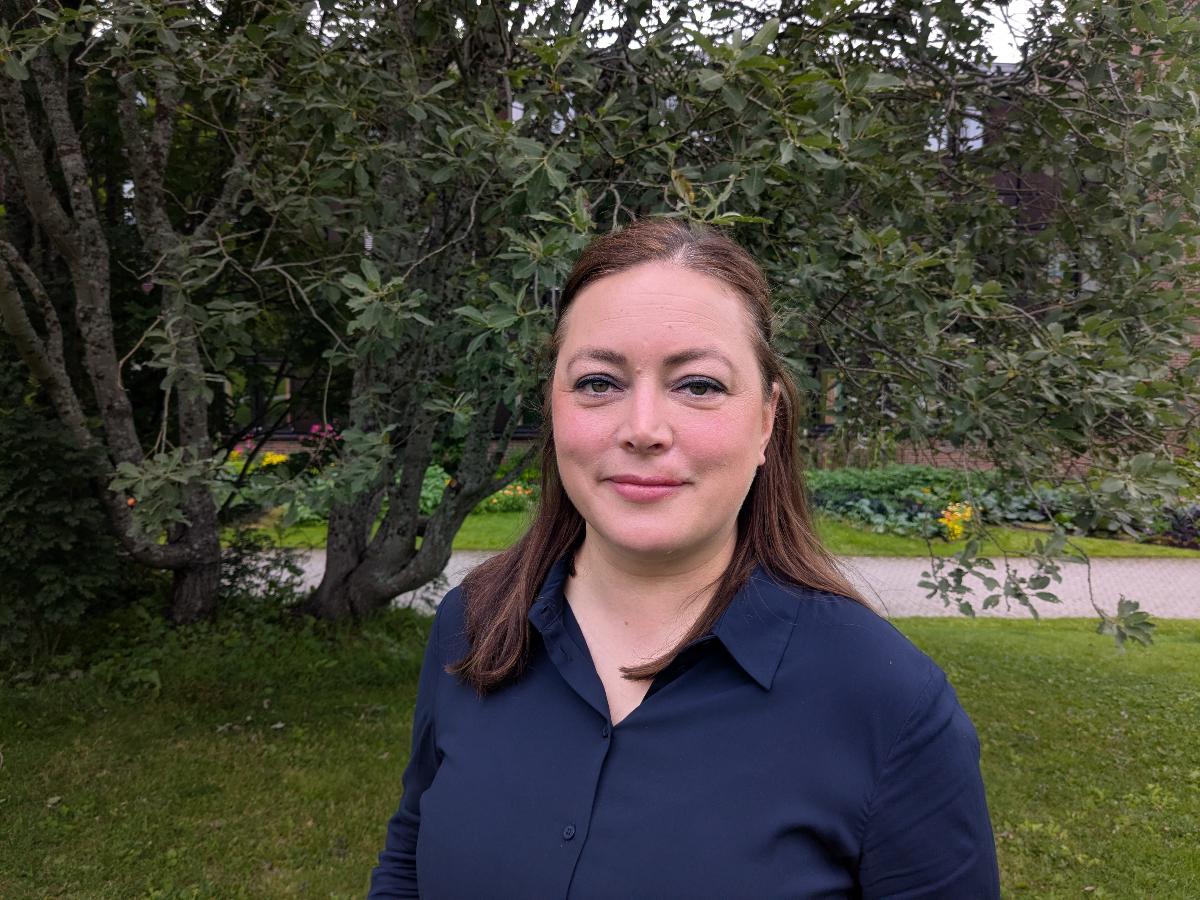 Portrettbilde av dame med m&oslash;rkt langt h&aring;r og bl&aring; skjorte som st&aring;r p&aring; en plen, foran et tre. 