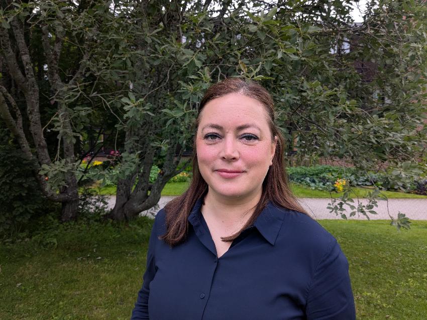 Portrettbilde av dame med mørkt langt hår og blå skjorte som står på en plen, foran et tre.