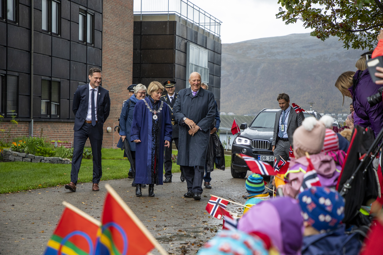 Kong Harald besøker UiT 2018.jpg King walking together with other leaders while kids are waving flags
