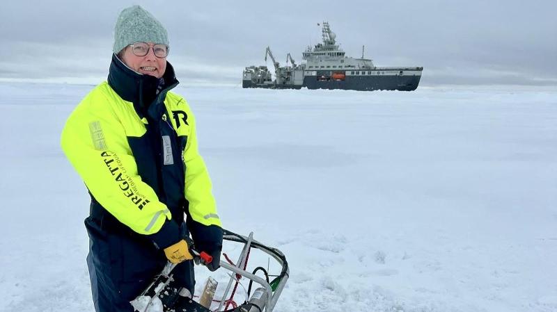 dame med vinterkl&aelig;r som holder et forsknings-instrument mens hun st&aring; p&aring; et isflak. forskningsskip i bakgrunnen.