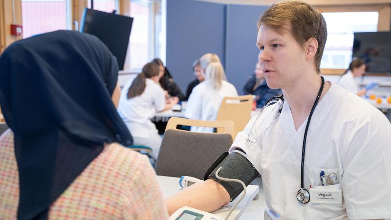 ung mann med helsefrakk og stetoskop rundt halsen og en dame med hijab med ryggen til i et klasserom