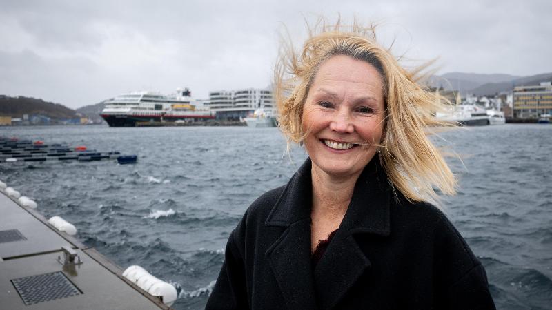 en kvinne i sterk vind p&aring; en kai. b&aring;ter og by i bakgrunnen